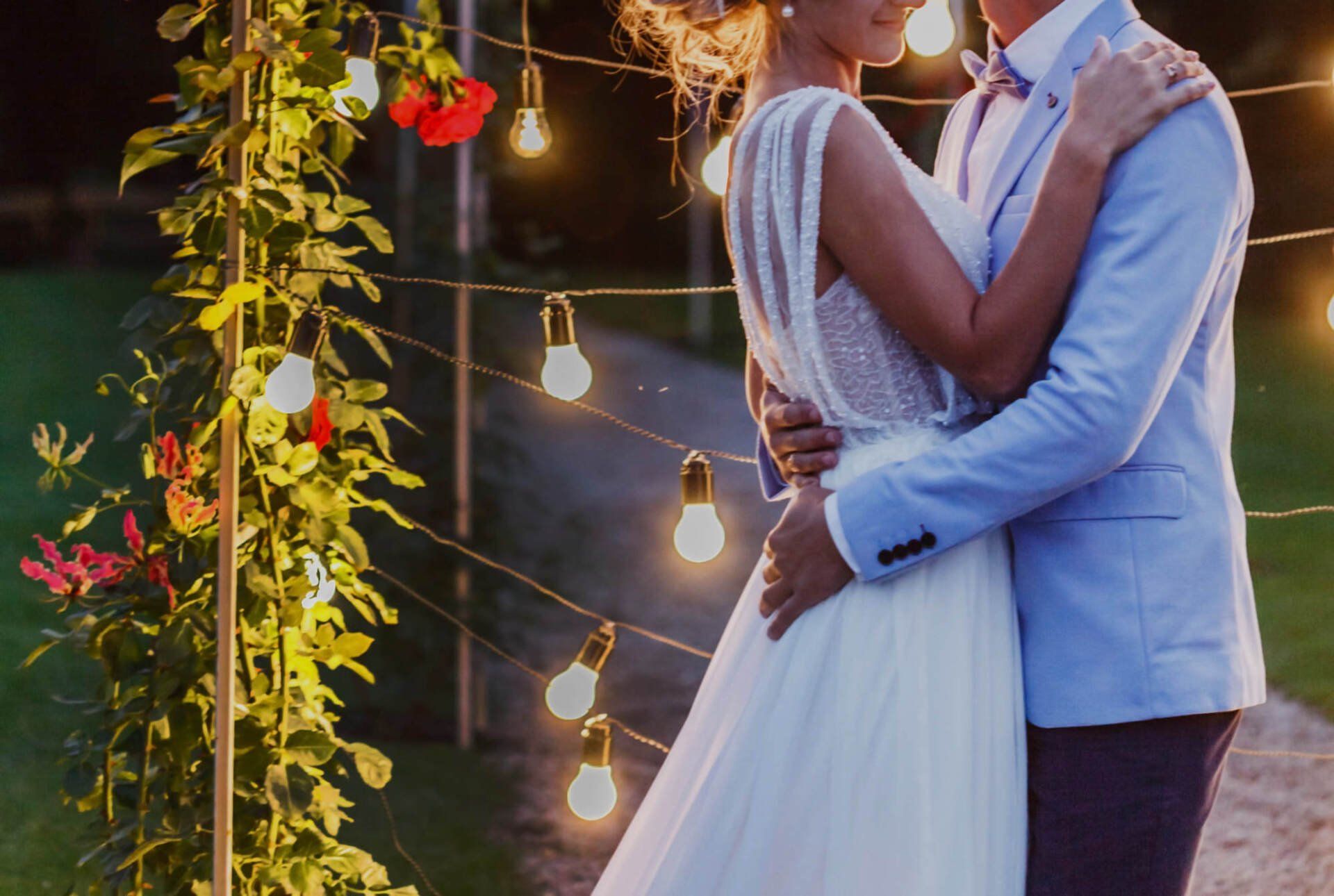 Couple embracing outdoors at dusk, with string lights and greenery.