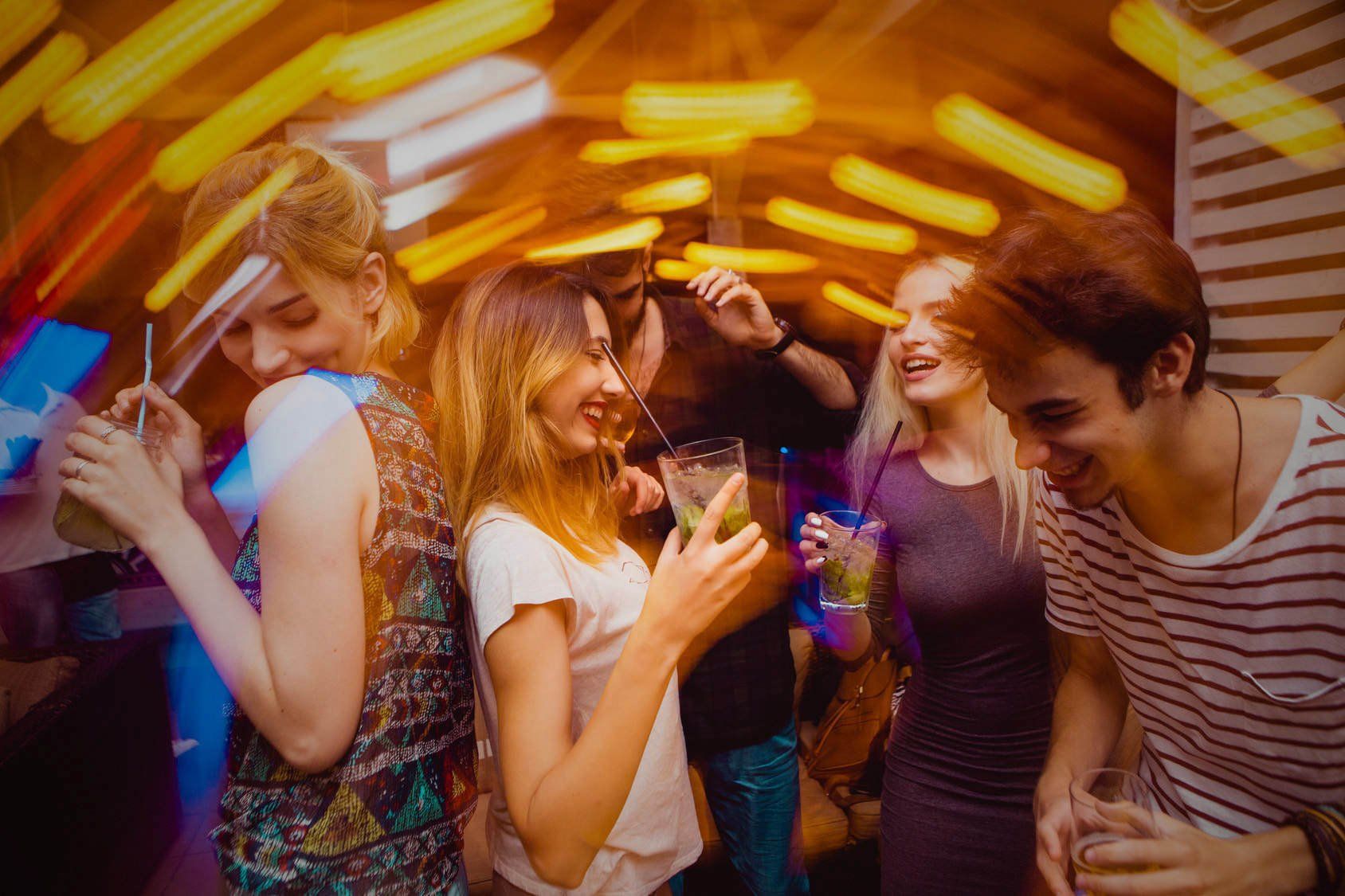 People dancing, holding drinks in a club with colorful, blurred lights.
