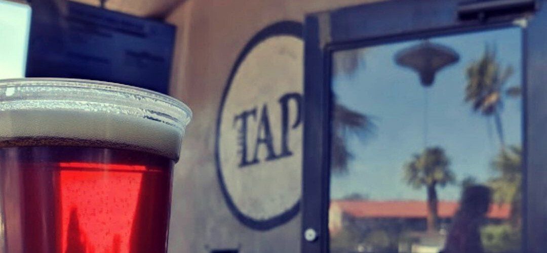 A glass of red beer with foam in front of a window reflecting palm trees and the word 