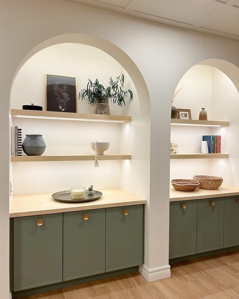 Deux alcôves cintrées avec armoires vert sauge, étagères en bois et objets décoratifs. Éclairage chaleureux.