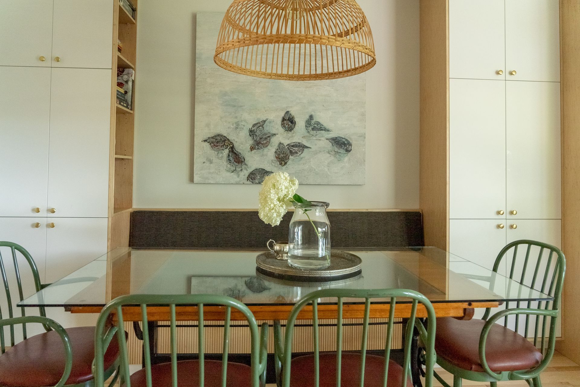 Salle à manger avec chaises vertes, table en verre, composition florale, art et plafonnier tissé.