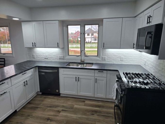 White kitchen with black countertops and appliances, wood flooring, and a window overlooking a yard.