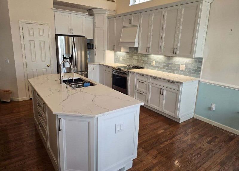 White kitchen with island, cabinets, and appliances. Wooden floors and light blue backsplash.