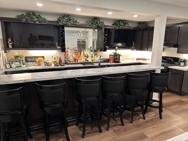Basement bar with dark wood cabinets, a long countertop with bar stools, and two TVs.