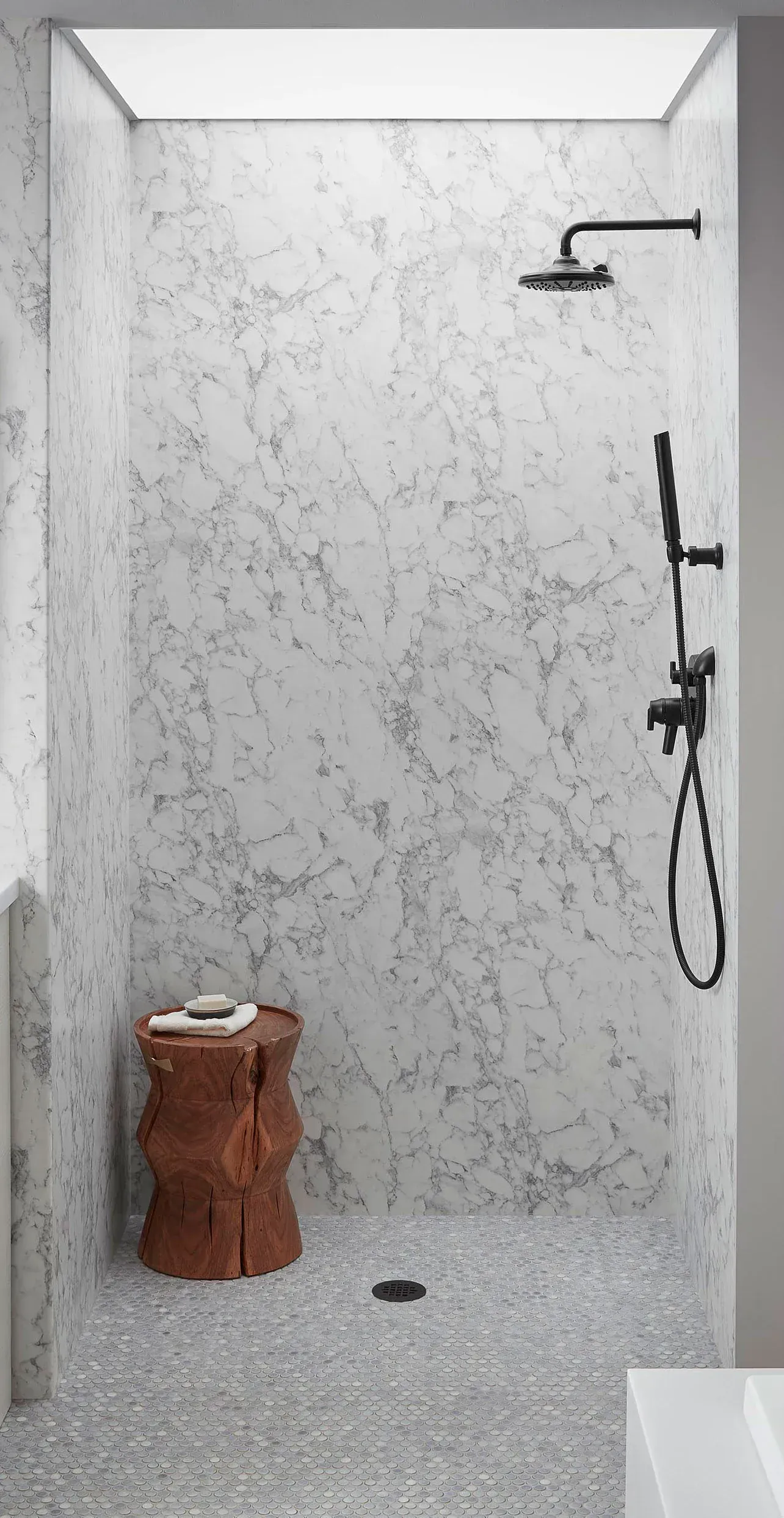 Shower with mottled gray walls and pebble floor, black fixtures, and a wooden stool.