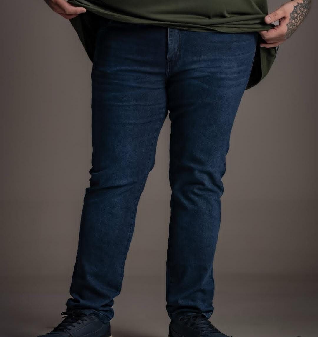 Persona que viste pantalones vaqueros azul oscuro y una camisa verde, de pie frente a un fondo gris.