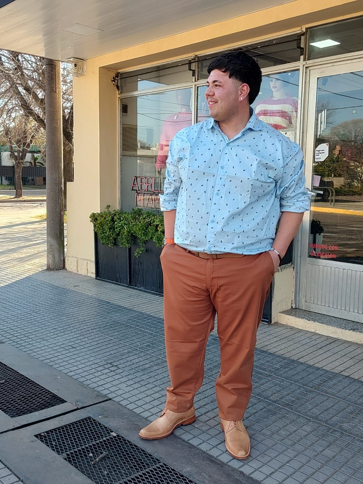 Hombre con camisa estampada azul y pantalones marrones de pie afuera de una tienda, sonriendo.