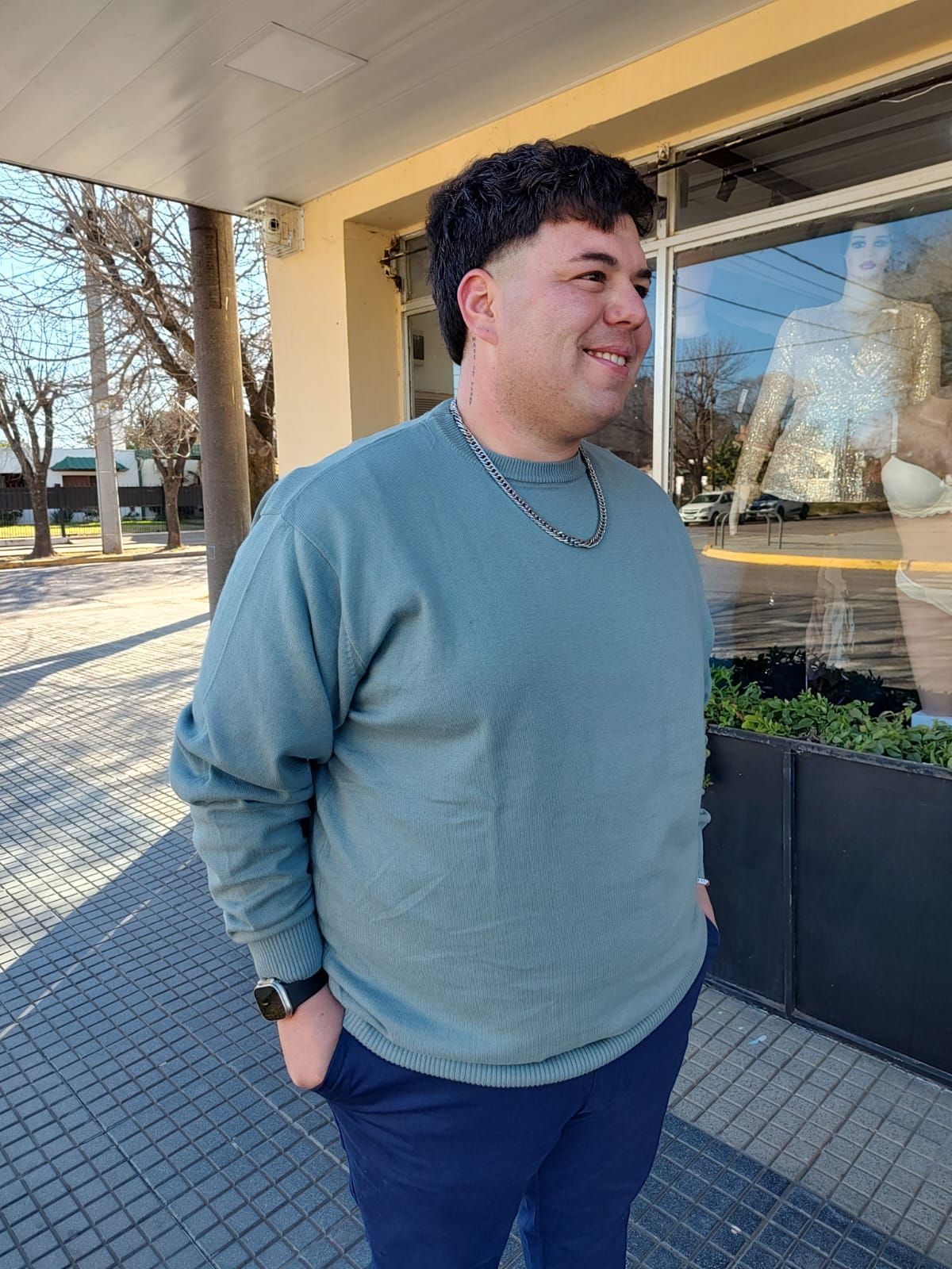 Hombre con suéter azul verdoso y pantalones azul marino sonriendo, con las manos en los bolsillos