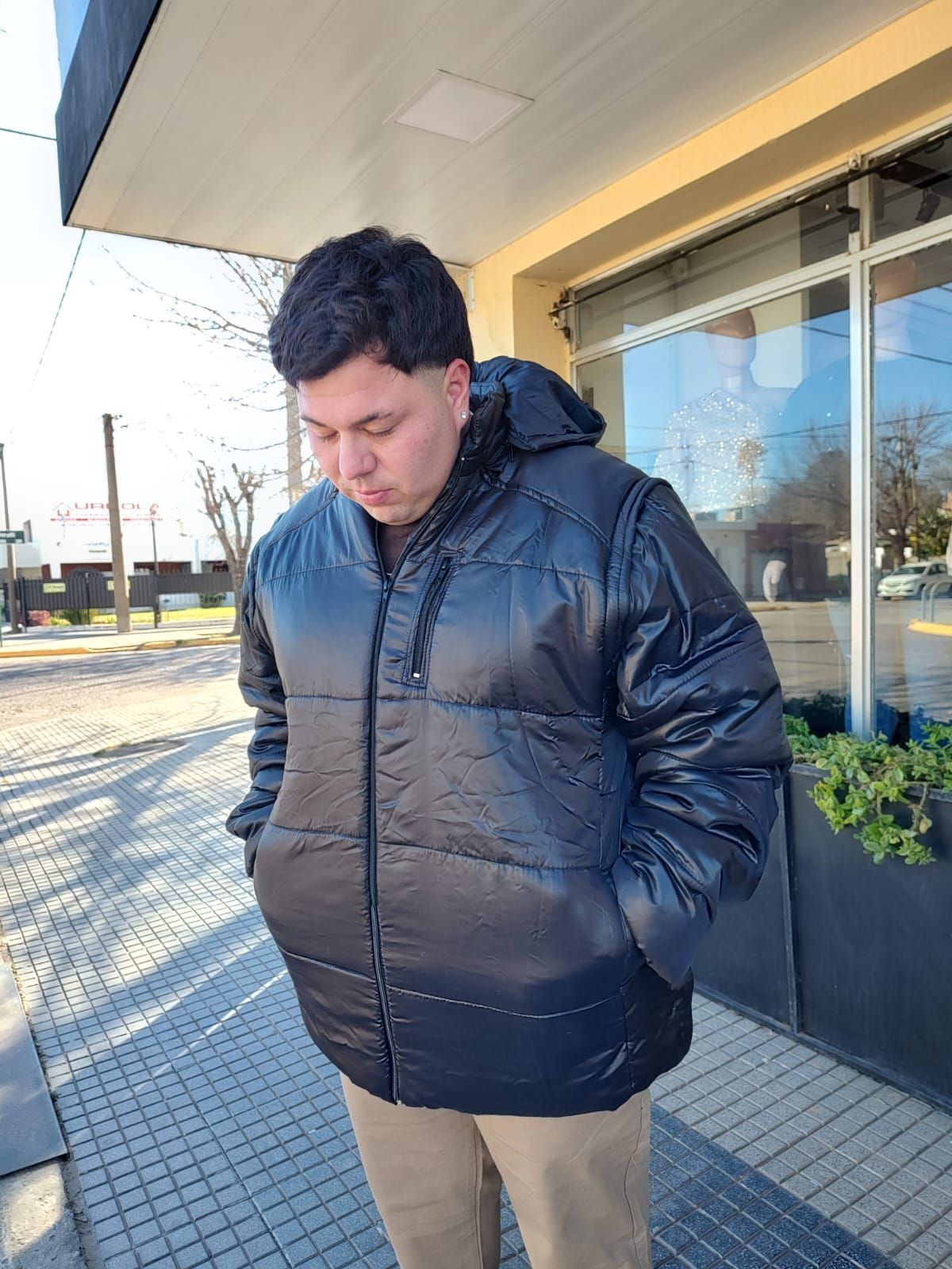 Hombre con una chaqueta negra inflada con capucha, de pie afuera de un edificio, con las manos en los bolsillos.