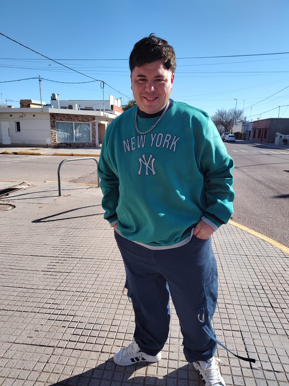 Hombre con sudadera verde azulado de Nueva York y pantalones deportivos azules de pie en una acera. Día soleado.