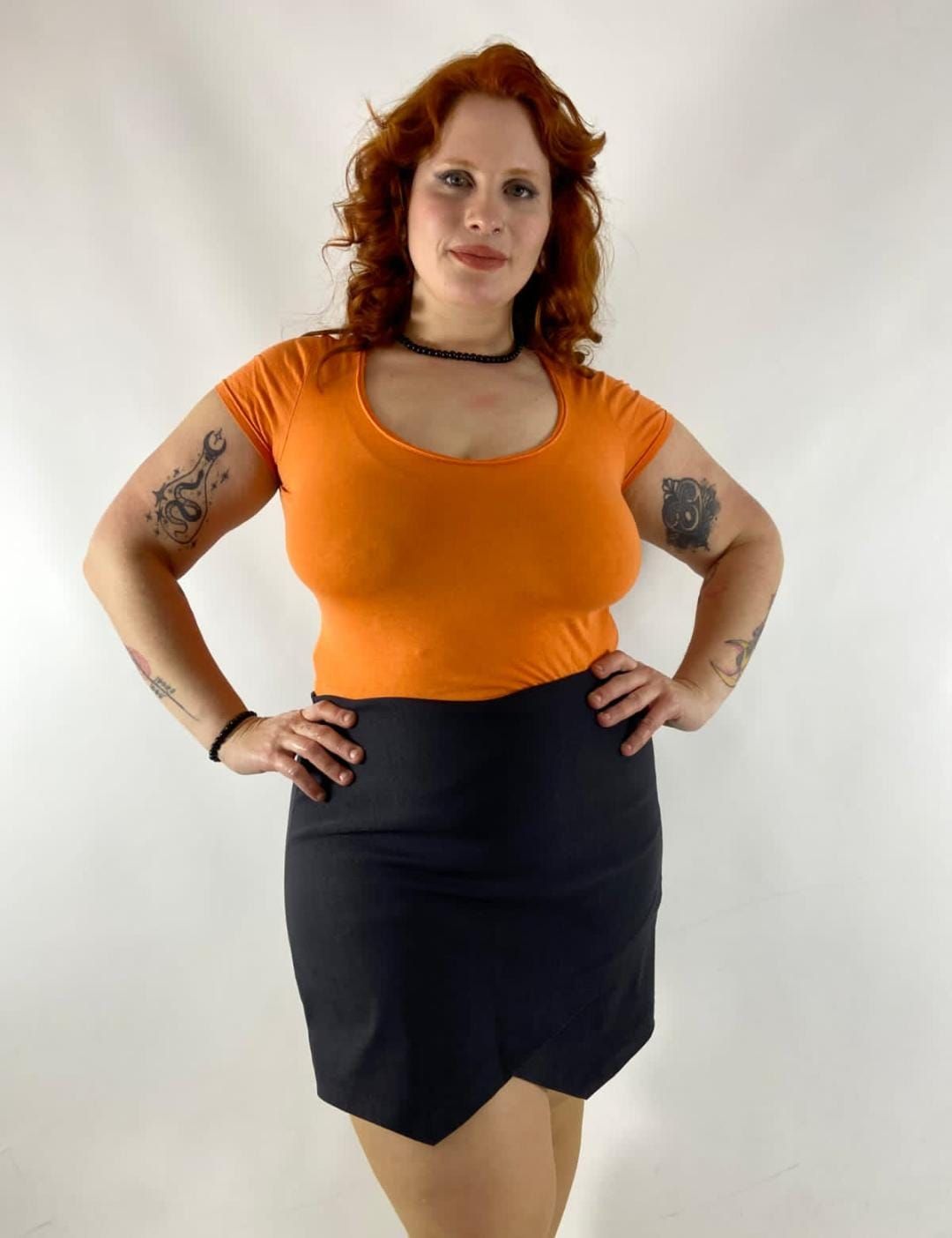 Mujer con camisa naranja y falda negra, con las manos en las caderas, sonriendo.