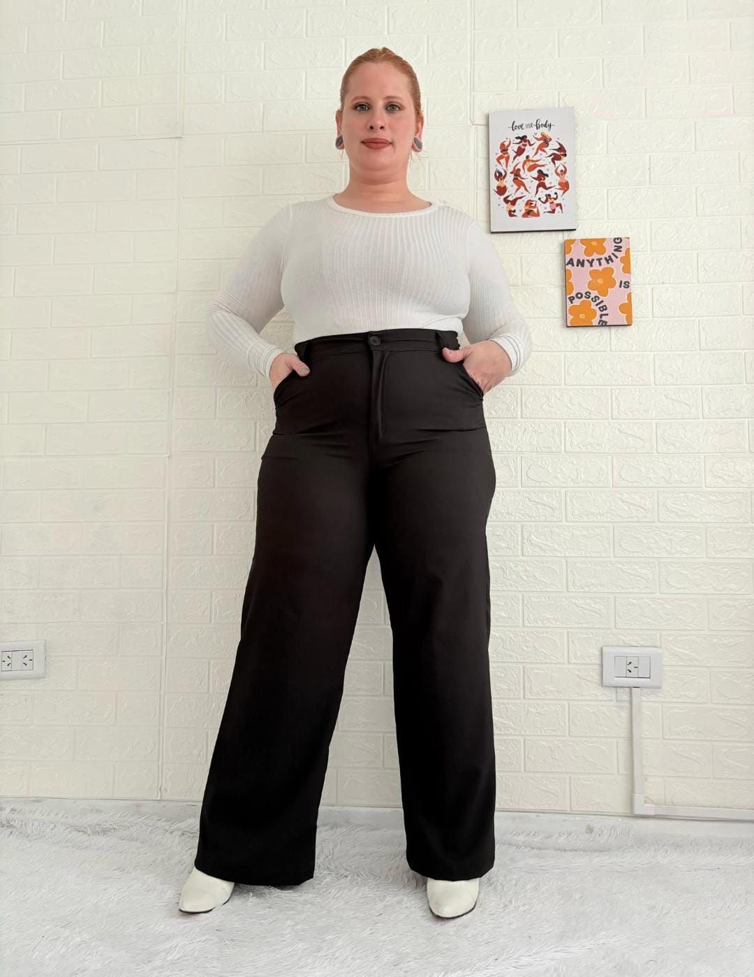 Mujer con suéter blanco y pantalones negros, con las manos en los bolsillos, posando frente a una pared de ladrillos blancos.