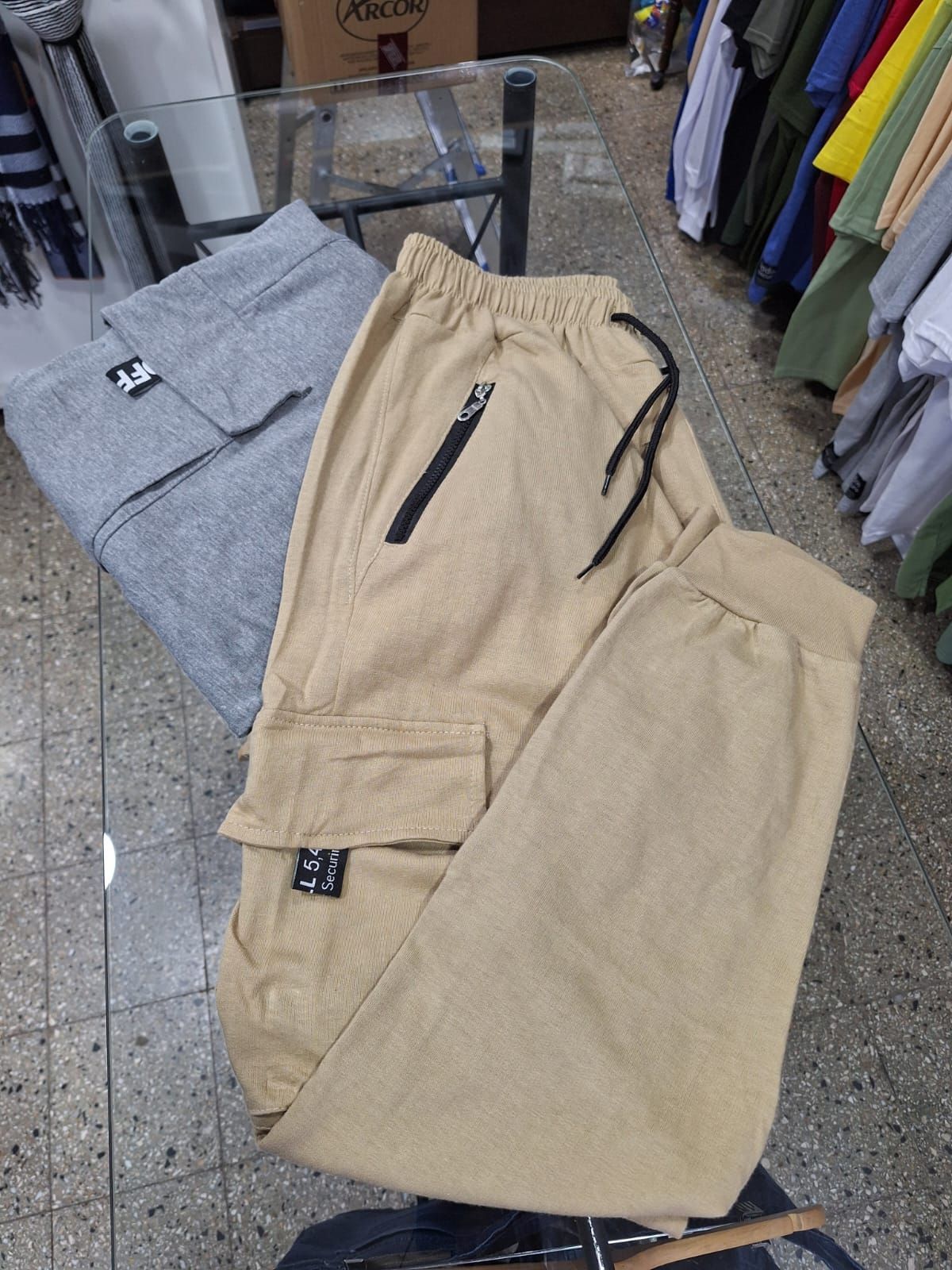 Pantalones cargo color caqui con cremallera negra y cordón en una vitrina de cristal; pantalones grises cerca.