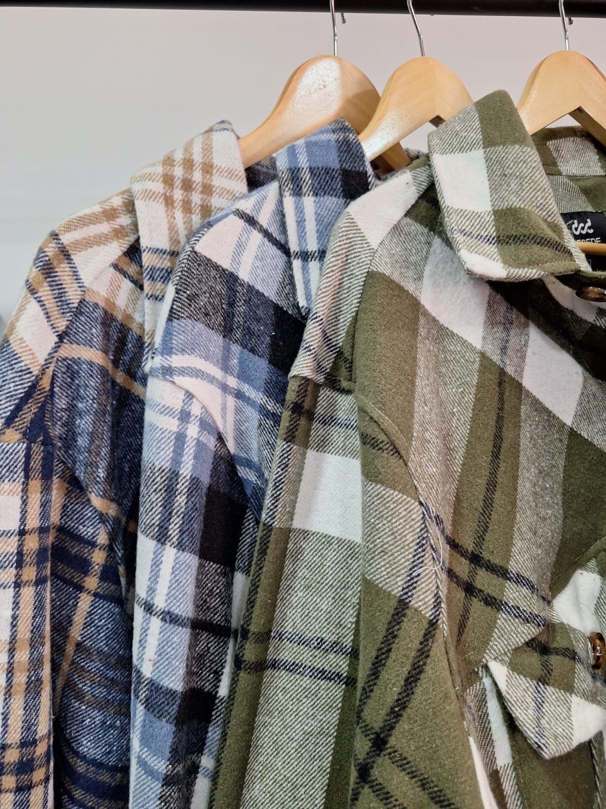 Tres camisas a cuadros en tonos tostados, azules y verdes colgadas en perchas de madera.