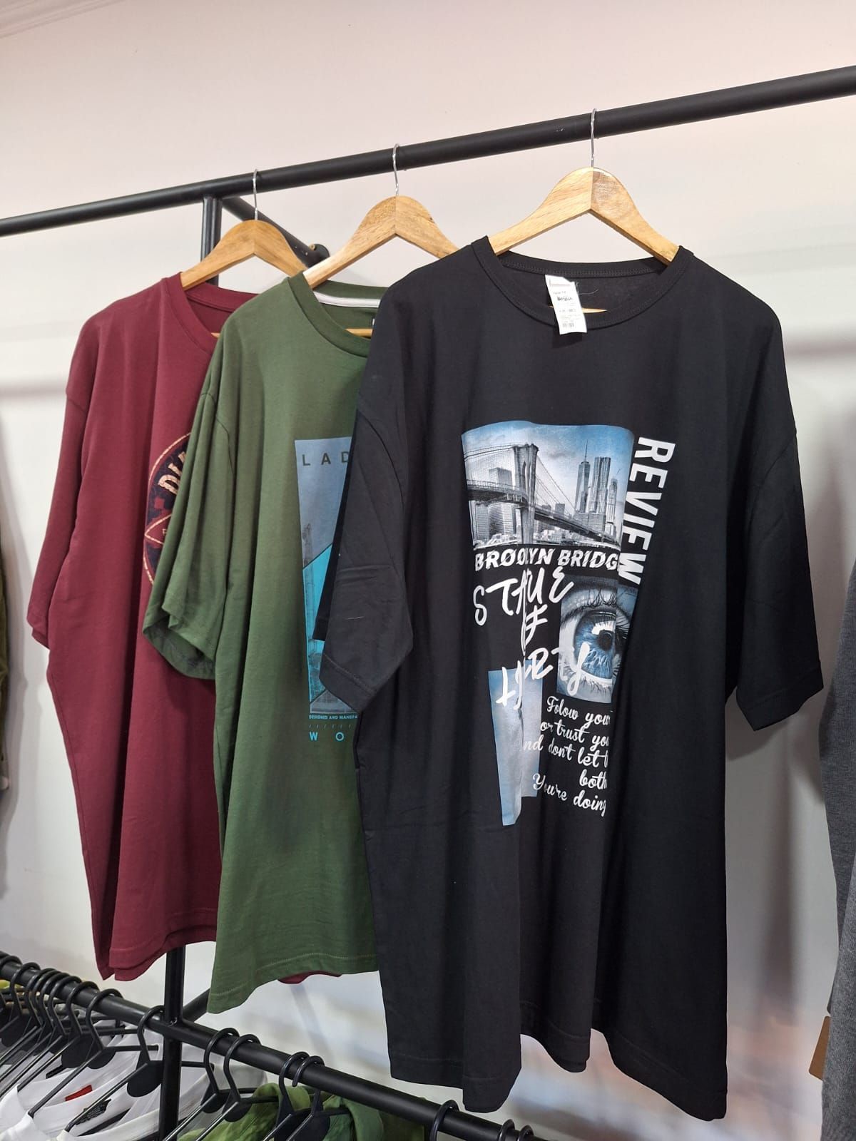 Tres camisetas extragrandes en perchas: granate, verde y negra. La camiseta negra tiene un puente.