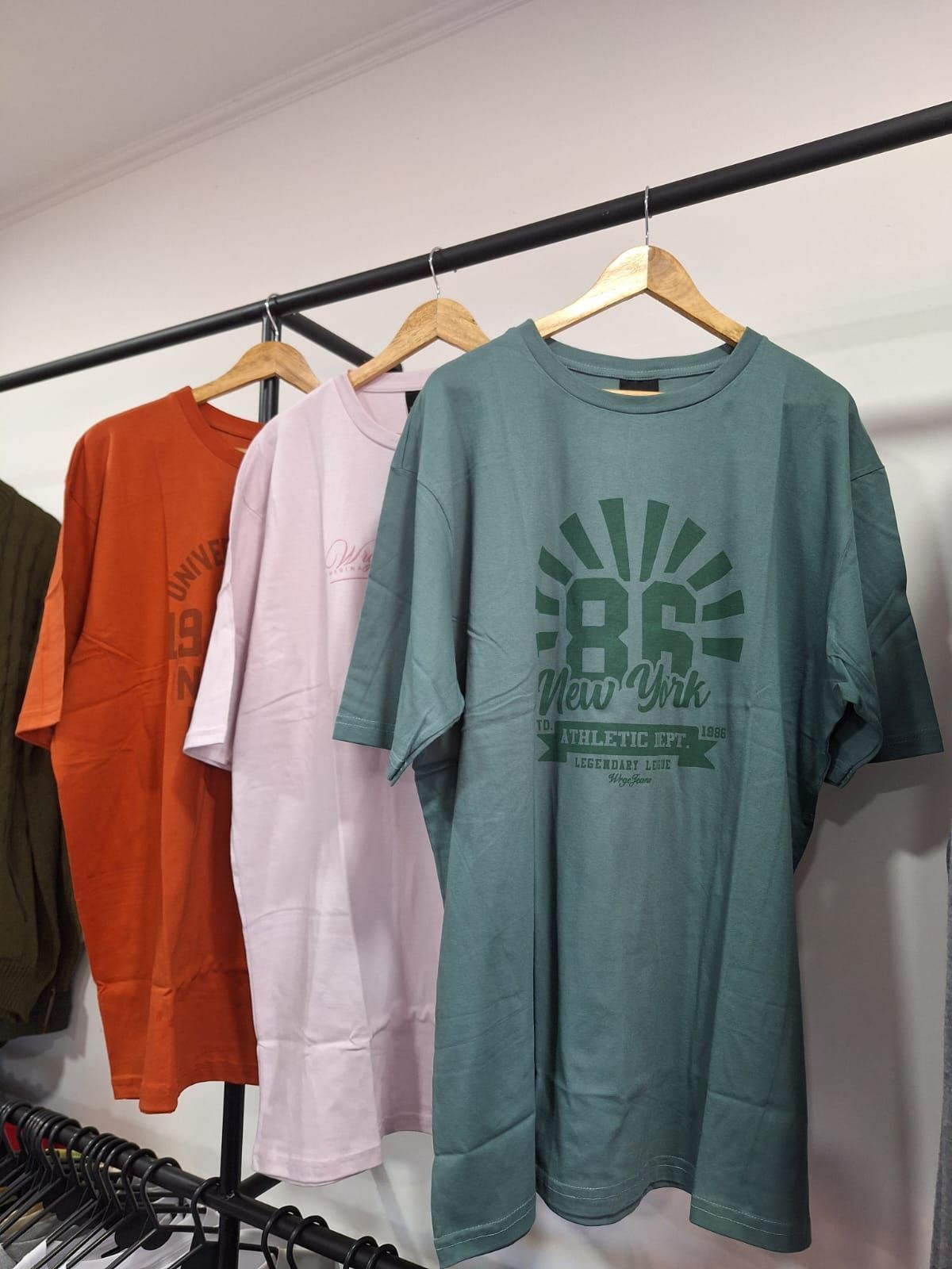 Tres camisetas gráficas de gran tamaño en tonos naranja, rosa y verde azulado colgadas en un perchero.