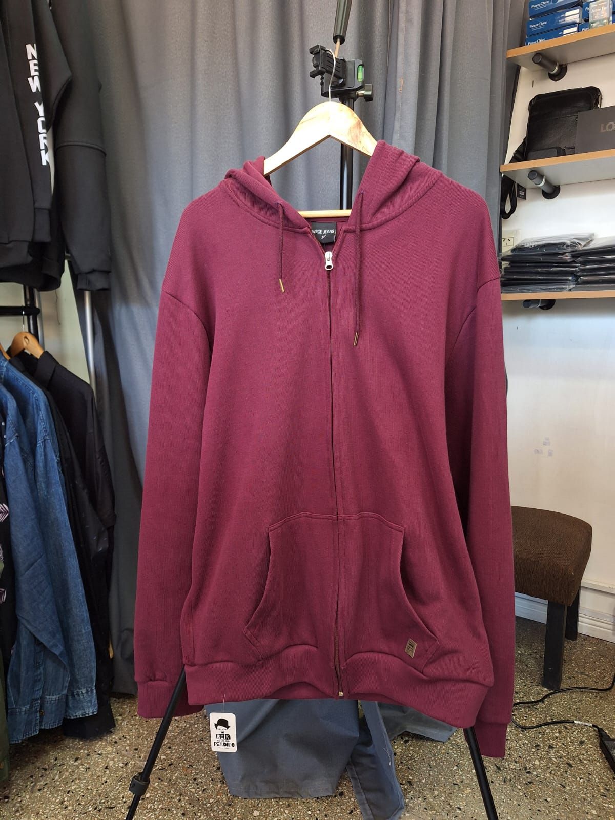Sudadera con capucha y cremallera de color burdeos colgada en una percha de madera en una tienda de ropa.