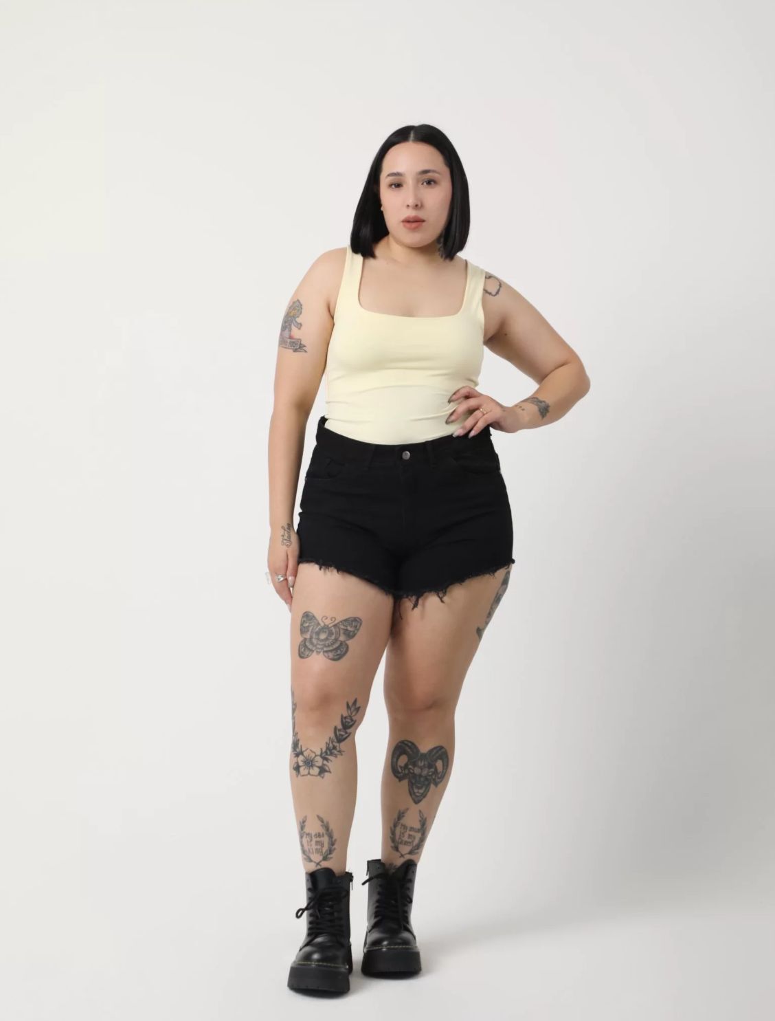Mujer con una camiseta amarilla y pantalones cortos negros con tatuajes, de pie frente a un fondo blanco.