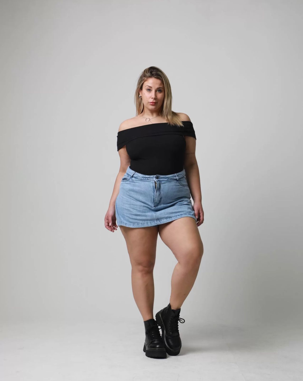 Mujer con top negro con hombros descubiertos, falda vaquera y botas negras posando en un estudio.