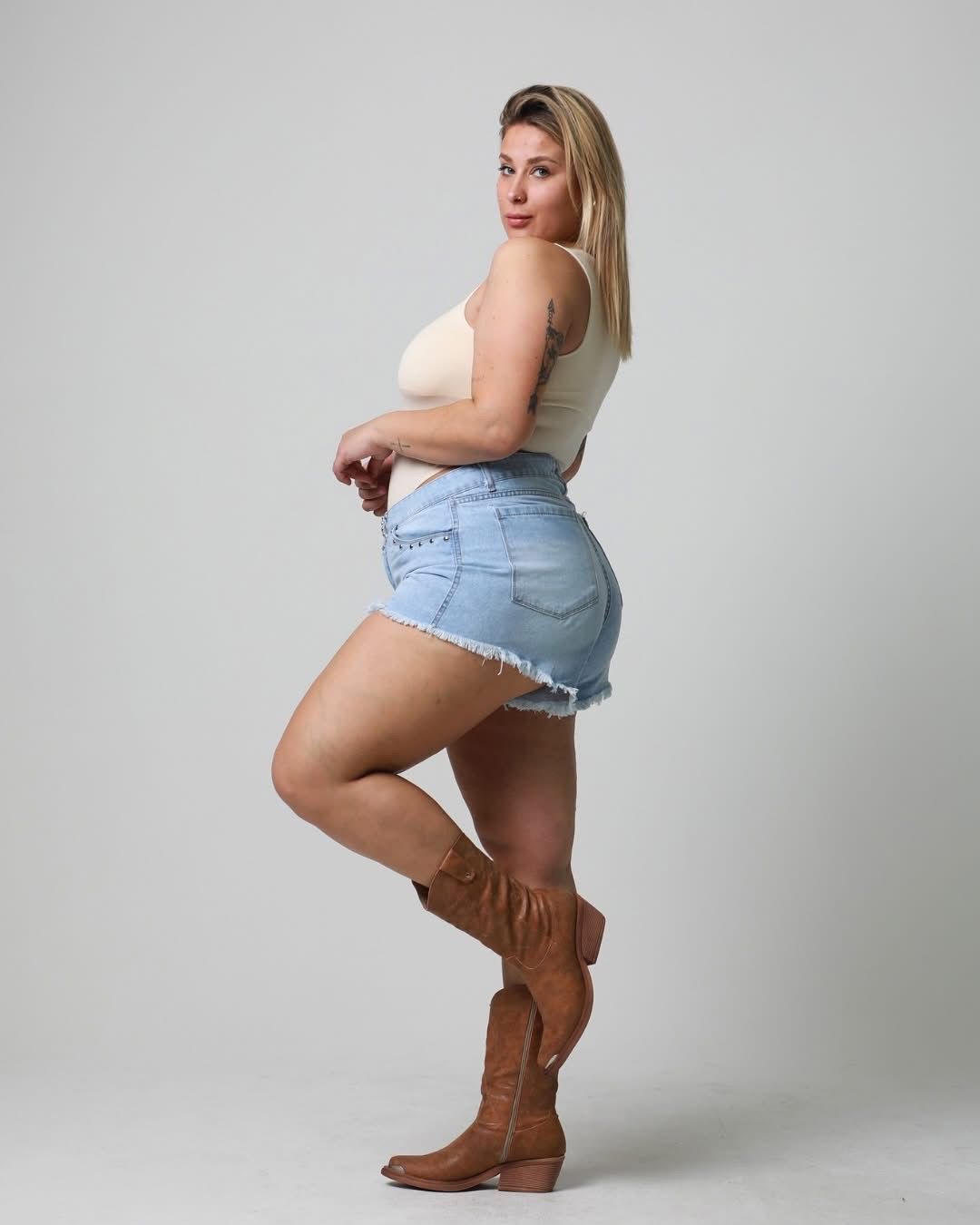 Una mujer con un traje de color claro, pantalones cortos de mezclilla y botas marrones posa frente a un fondo blanco.