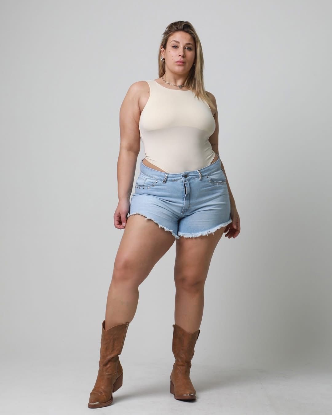 Una mujer con un traje de color claro, pantalones cortos de mezclilla y botas marrones posa frente a un fondo blanco.