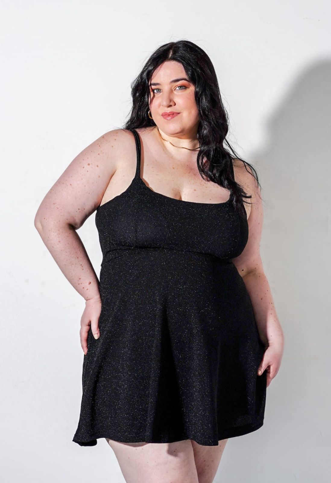 Mujer con un vestido negro brillante, de pie con las manos en las caderas, sonriendo, contra un fondo blanco.