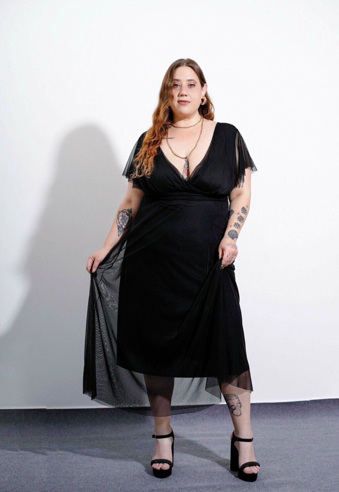 Mujer con un vestido negro con mangas transparentes, tacones negros, sosteniendo su vestido, de pie frente a un fondo blanco.
