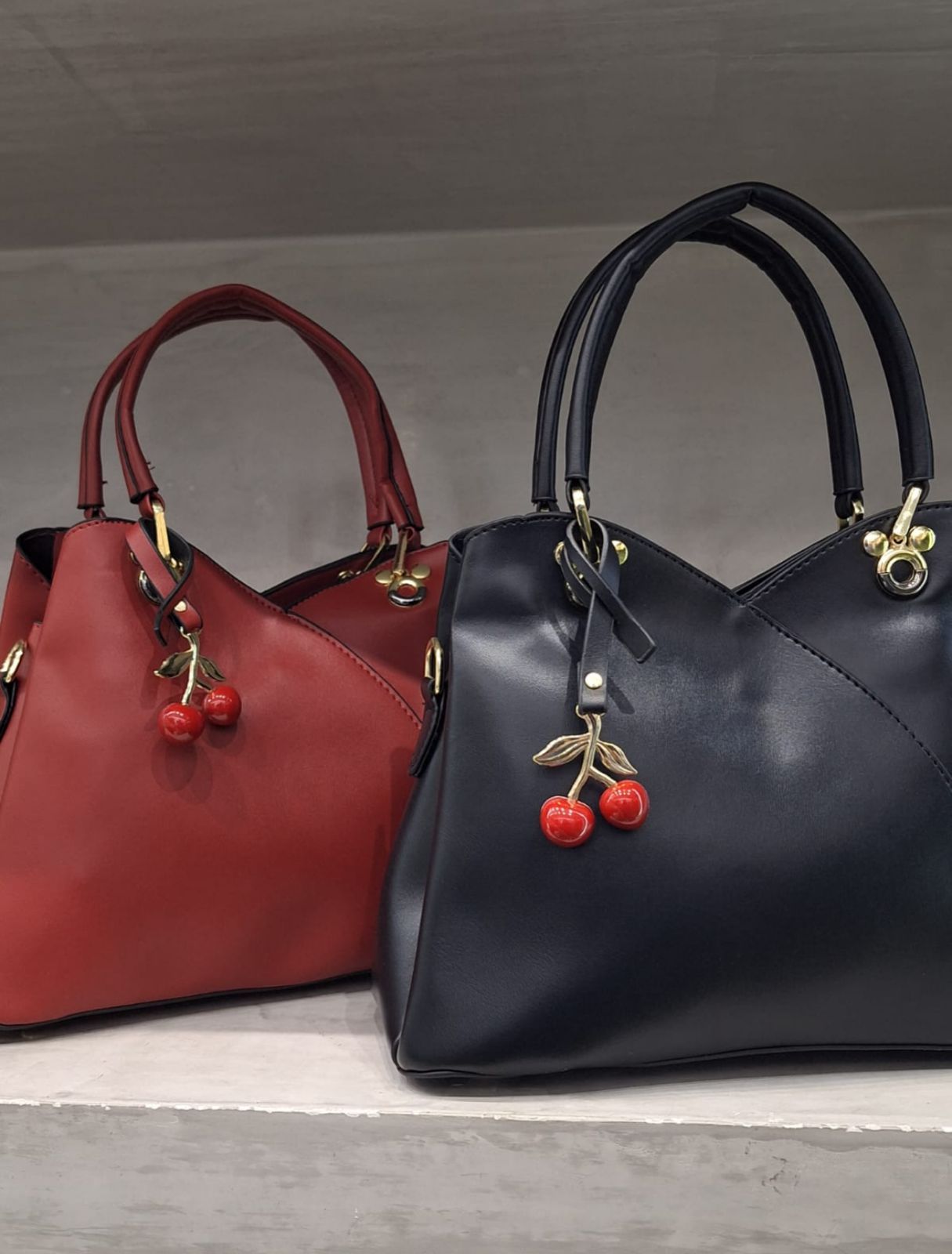 Dos bolsos de piel, uno rojo y otro negro, con charms en forma de cereza colgando de las asas.