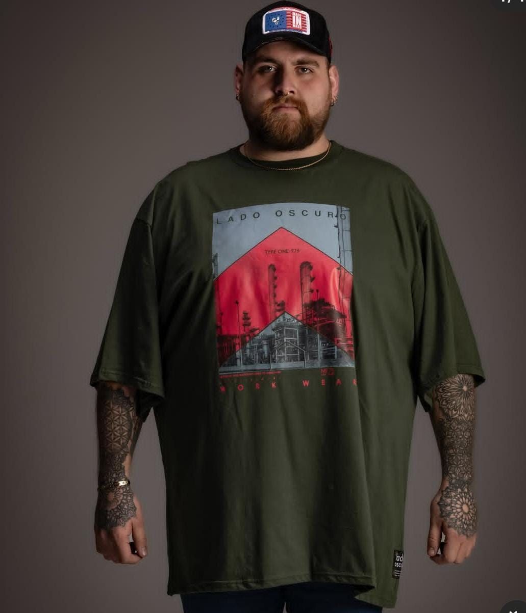 Hombre con barba y tatuajes, con camiseta y gorra verdes con el dibujo de un edificio. Fondo gris.