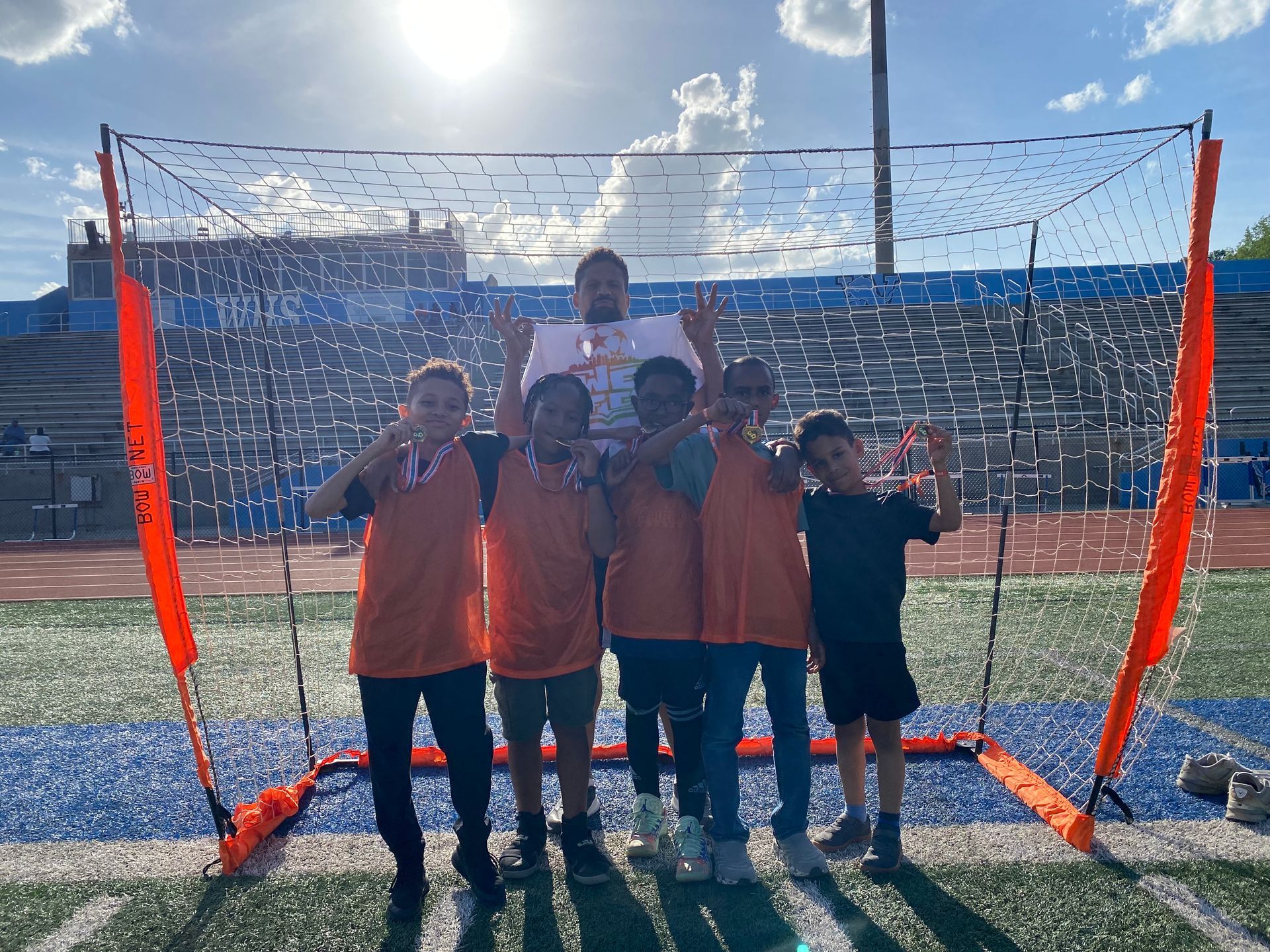 Five children pose in orange jerseys in front of a soccer goal on a sunny field.