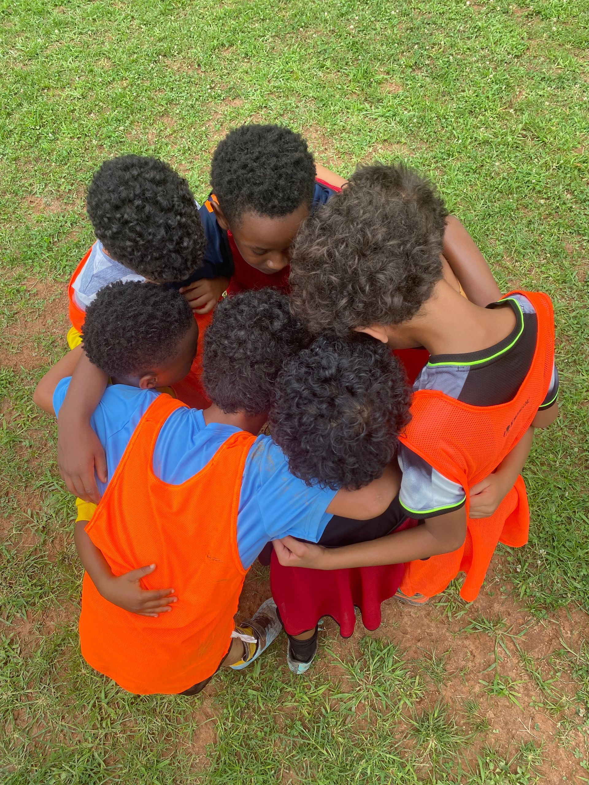 Five children in orange vests huddle together on grass, arms around each other.