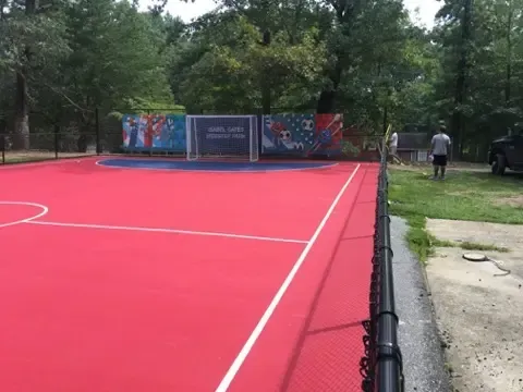 Bright red outdoor court with white lines, surrounded by trees and a fence, with a person walking nearby.