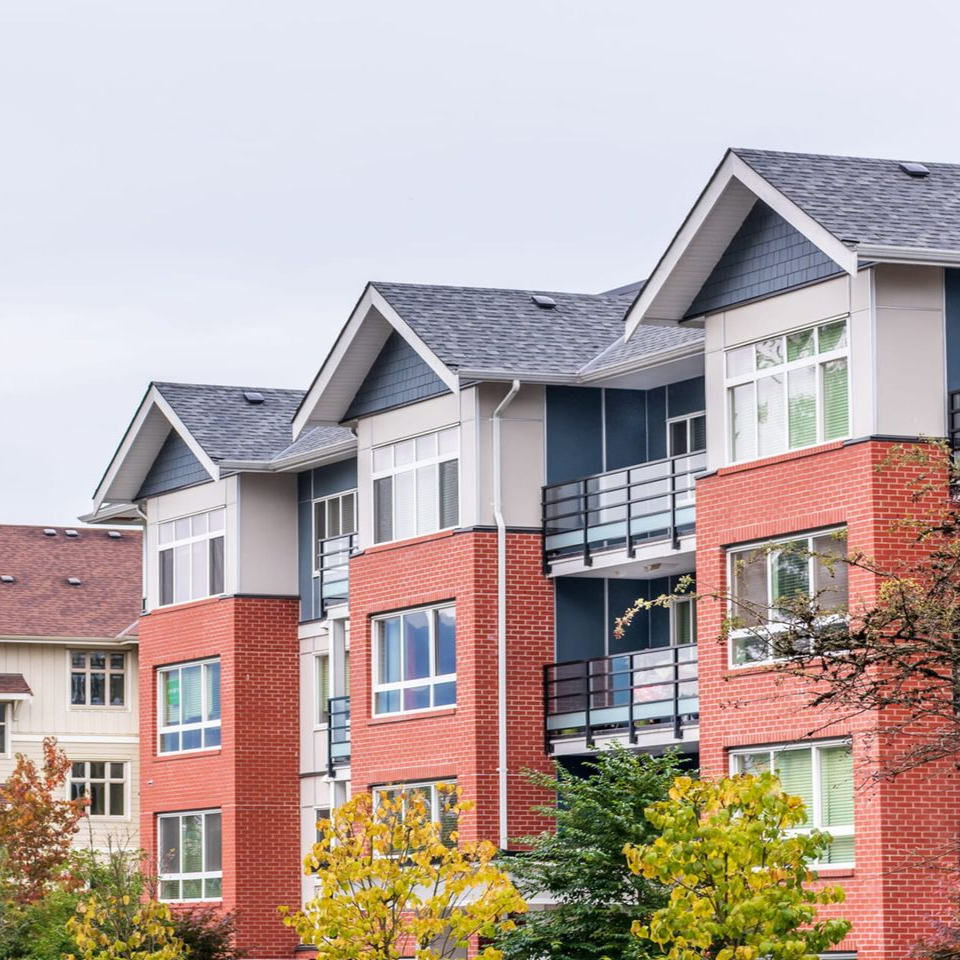 Modern apartment complex with red brick facades, dark blue trim, and several balconies under an overcast sky.
