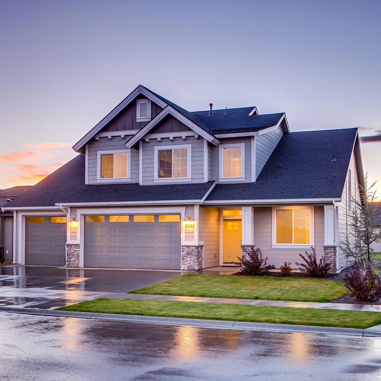 A multi-story suburban house with a two-car garage at twilight, featuring grey siding and a dark roof under a soft sky.
