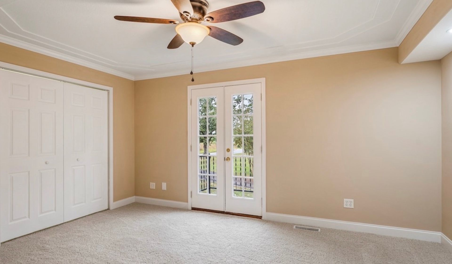 Empty beige bedroom with carpet, ceiling fan, closet doors, and French doors to outside.