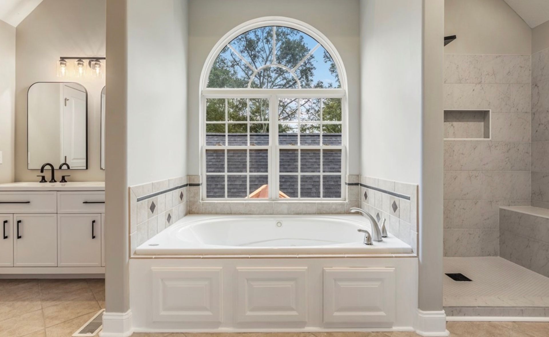 Bright bathroom with a white soaking tub under an arched window and adjacent walk-in shower