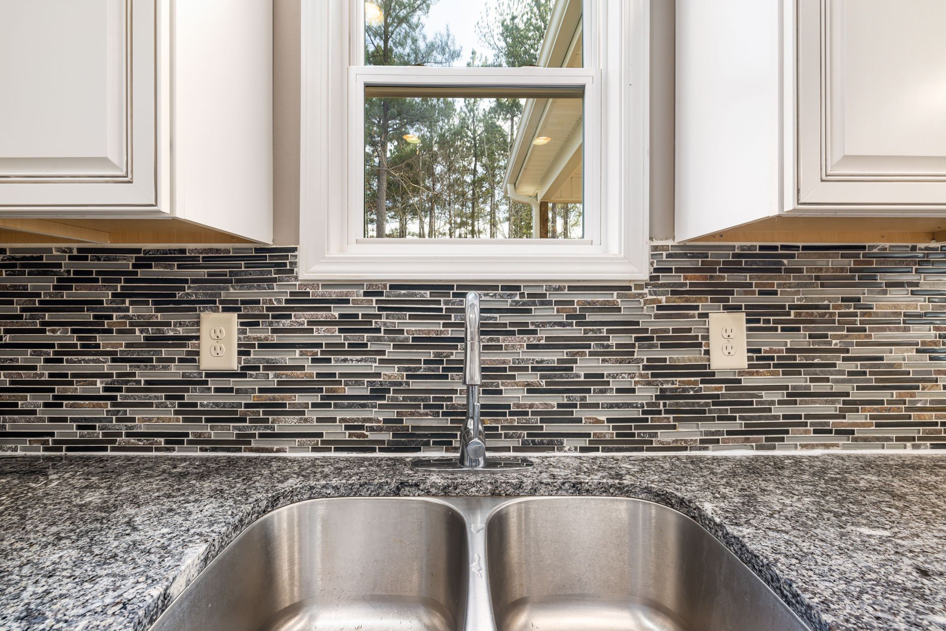 Kitchen sink beneath window with white cabinets, gray granite counters, and dark mosaic tile backsplash