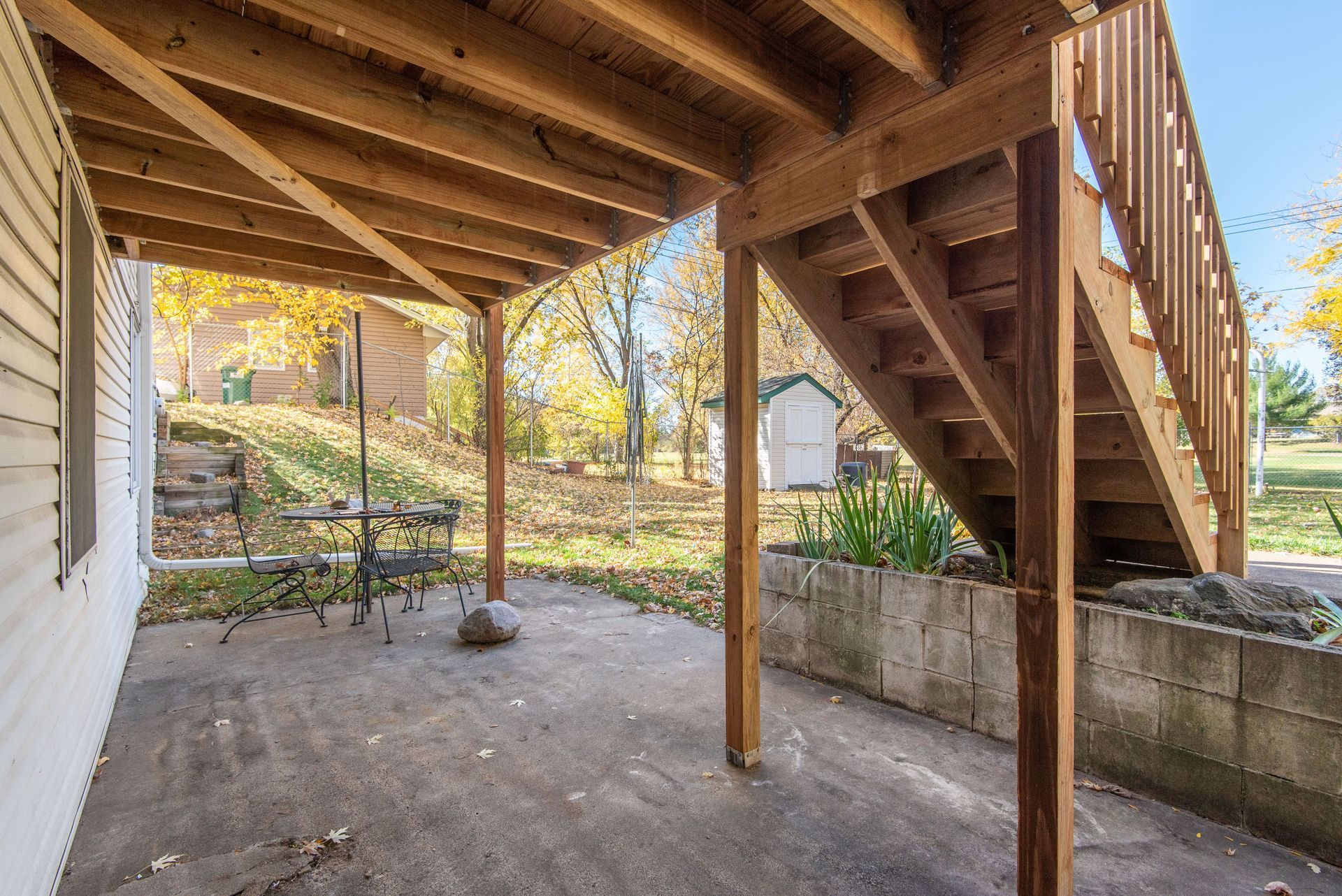 Under-deck concrete patio with wooden stairs, support posts, and a backyard view.