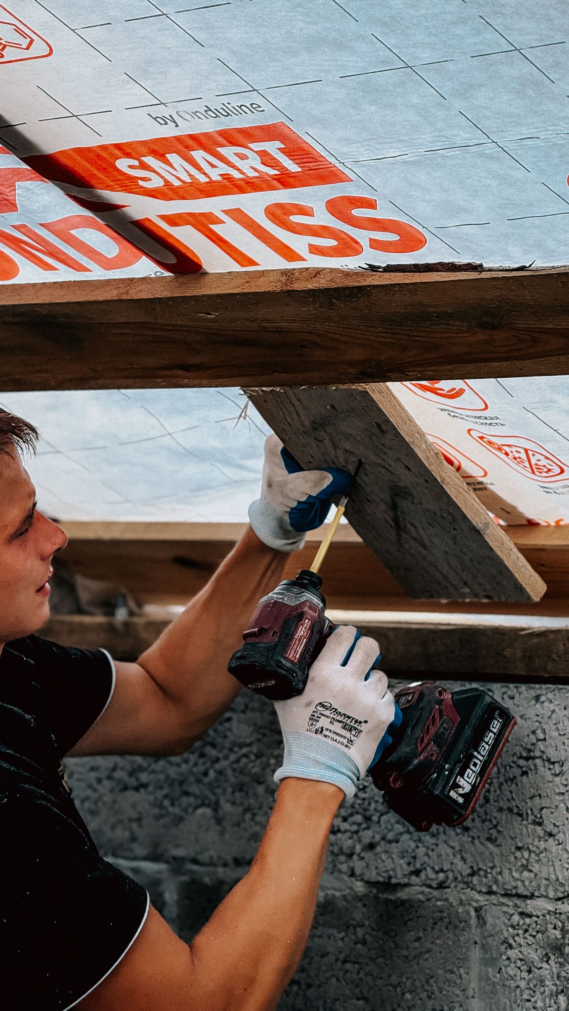 Worker in white gloves drilling wood under a roof with orange insulation wrap above.