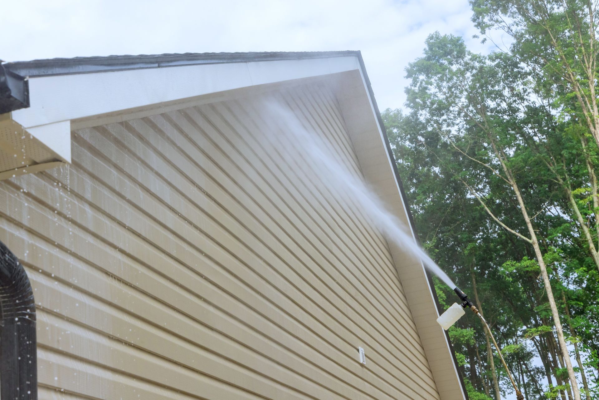 Person spraying water on a beige house siding and roof eave outdoors with trees in the background