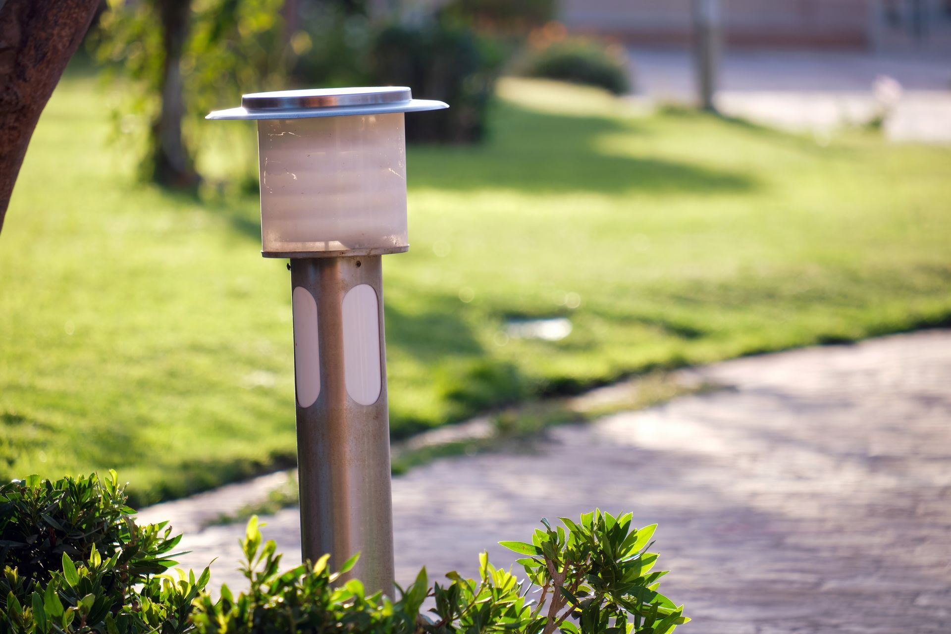 Metal garden light along a sunlit walkway beside trimmed bushes and green lawn