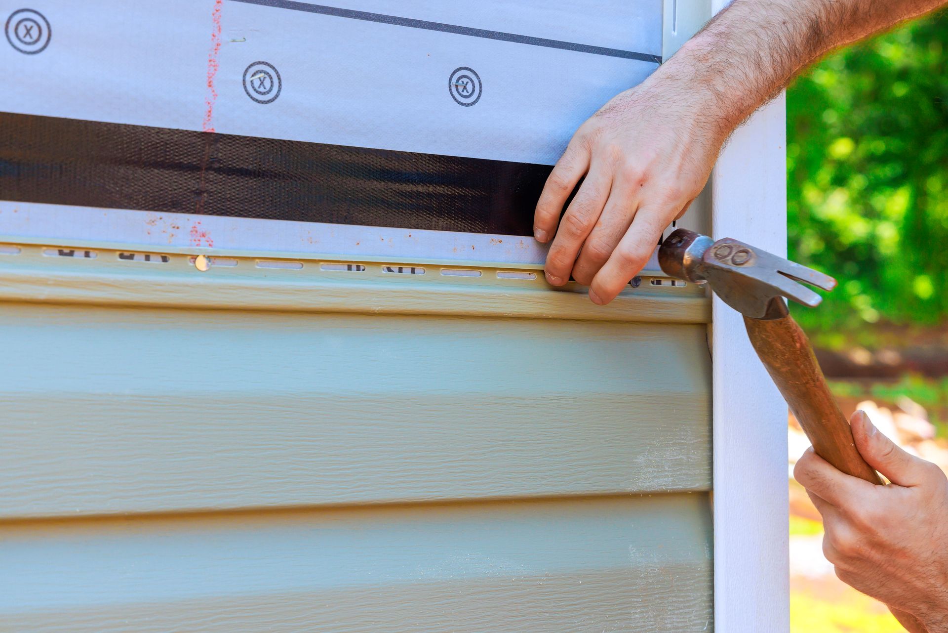 Hands use a crowbar to pry off beige siding on a house exterior.