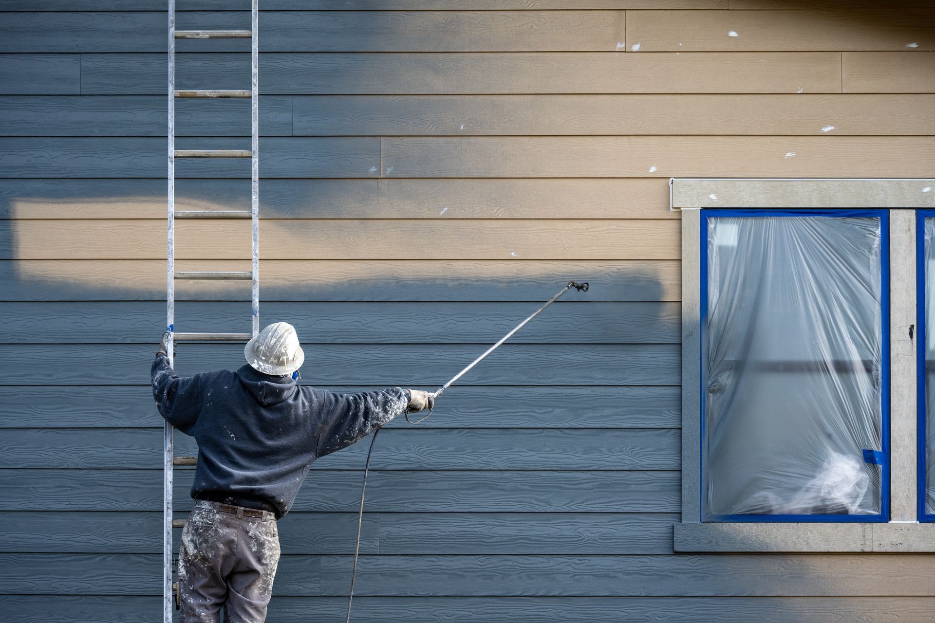 Person on a ladder painting a gray house exterior near a blue-framed window.