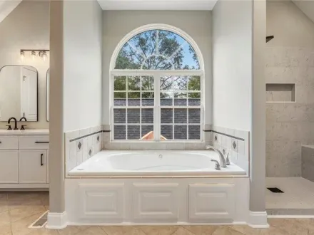 Bright bathroom with a white soaking tub under an arched window and tiled shower on the right.
