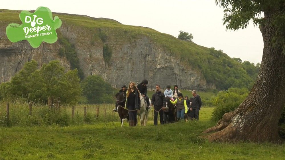 People on horseback and walking, on a grassy field near a rock face and trees.