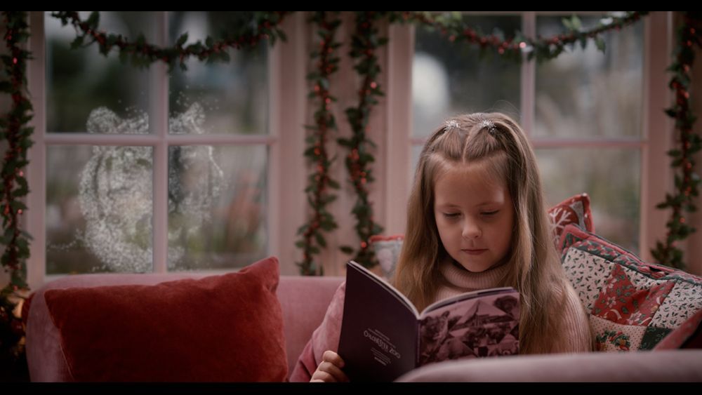 Girl reading a book by a window, with decorations. Two ghostly figures visible outside the window.