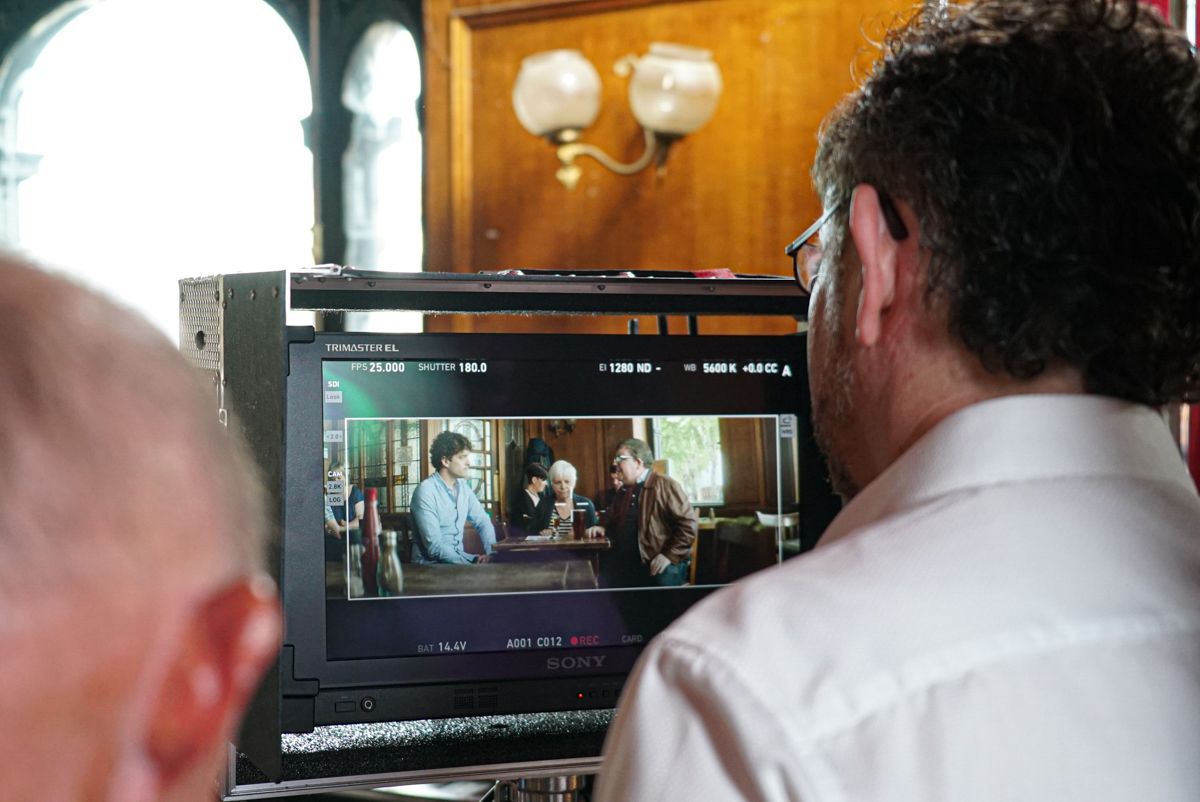 A monitor displays a scene of people at a table, viewed by a crew member.