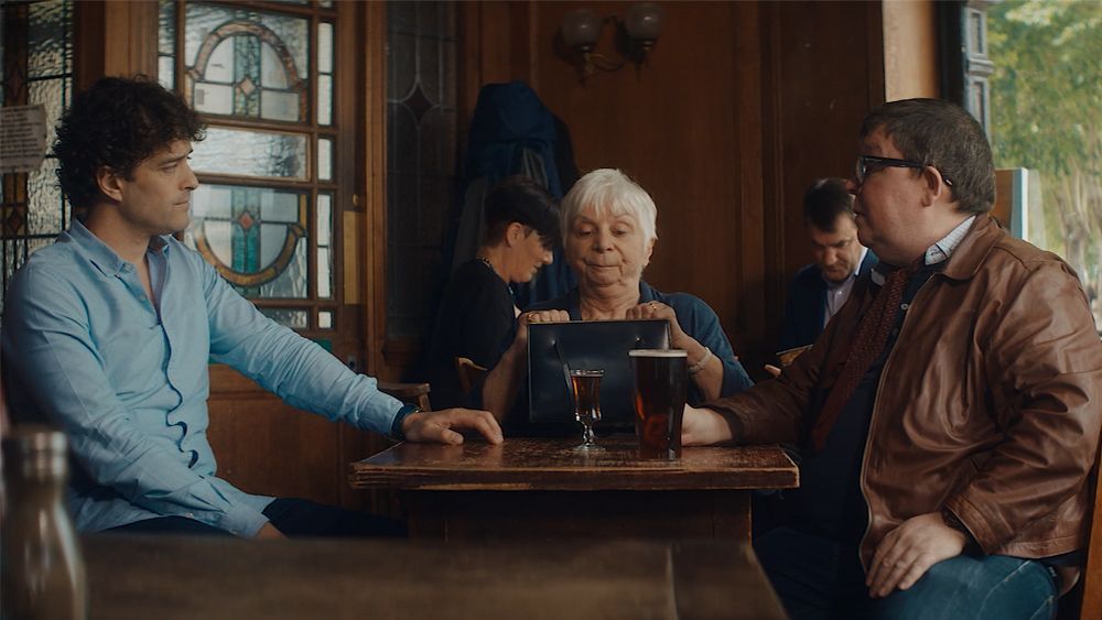 Three people at a pub table looking at a tablet, others in background. Man on left, woman in the middle, man on the right.