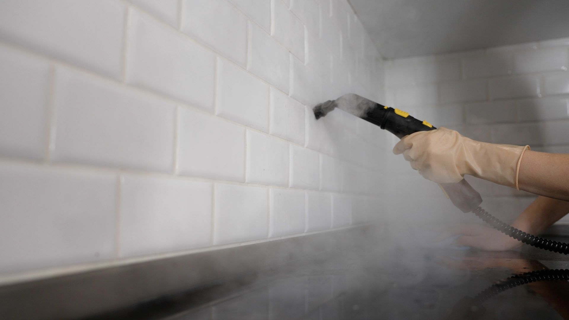 Person steaming a white tiled wall with a handheld steam cleaner, creating a cloud of steam.