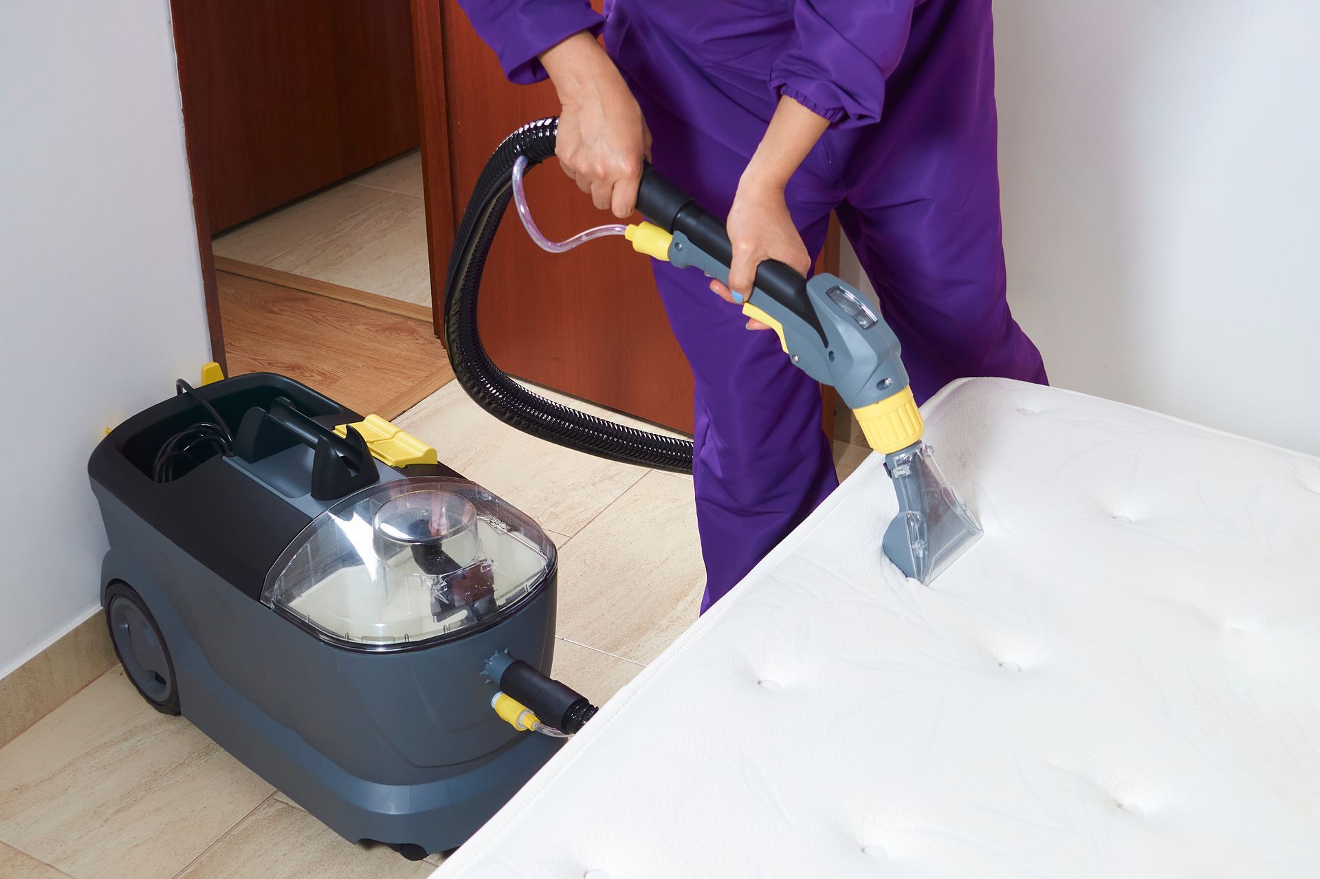 Person in purple cleans a white mattress with a handheld steam vacuum.